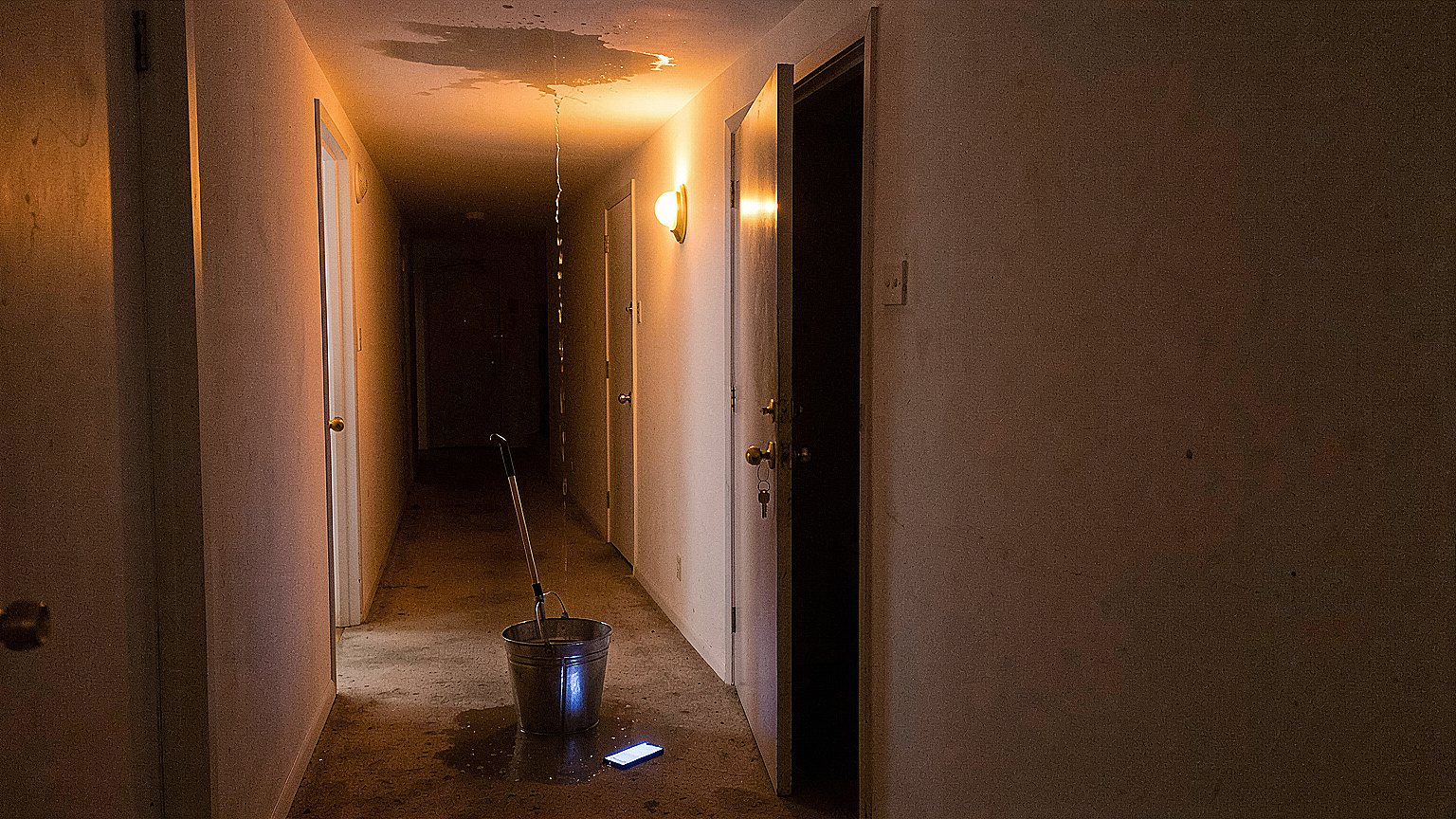 A dim apartment hallway at night with water dripping from a stained ceiling toward a steel mop bucket, a lit smartphone on the carpet, and keys hanging from the handle of an open door